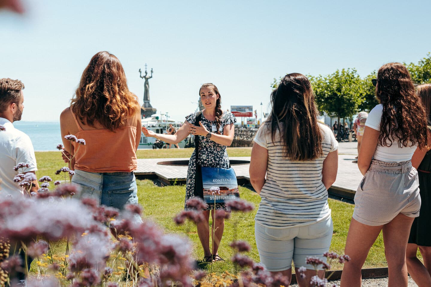 A group listens intently to the city guide during a city tour at the port of Constance. The Imperia statue can be seen in the background.