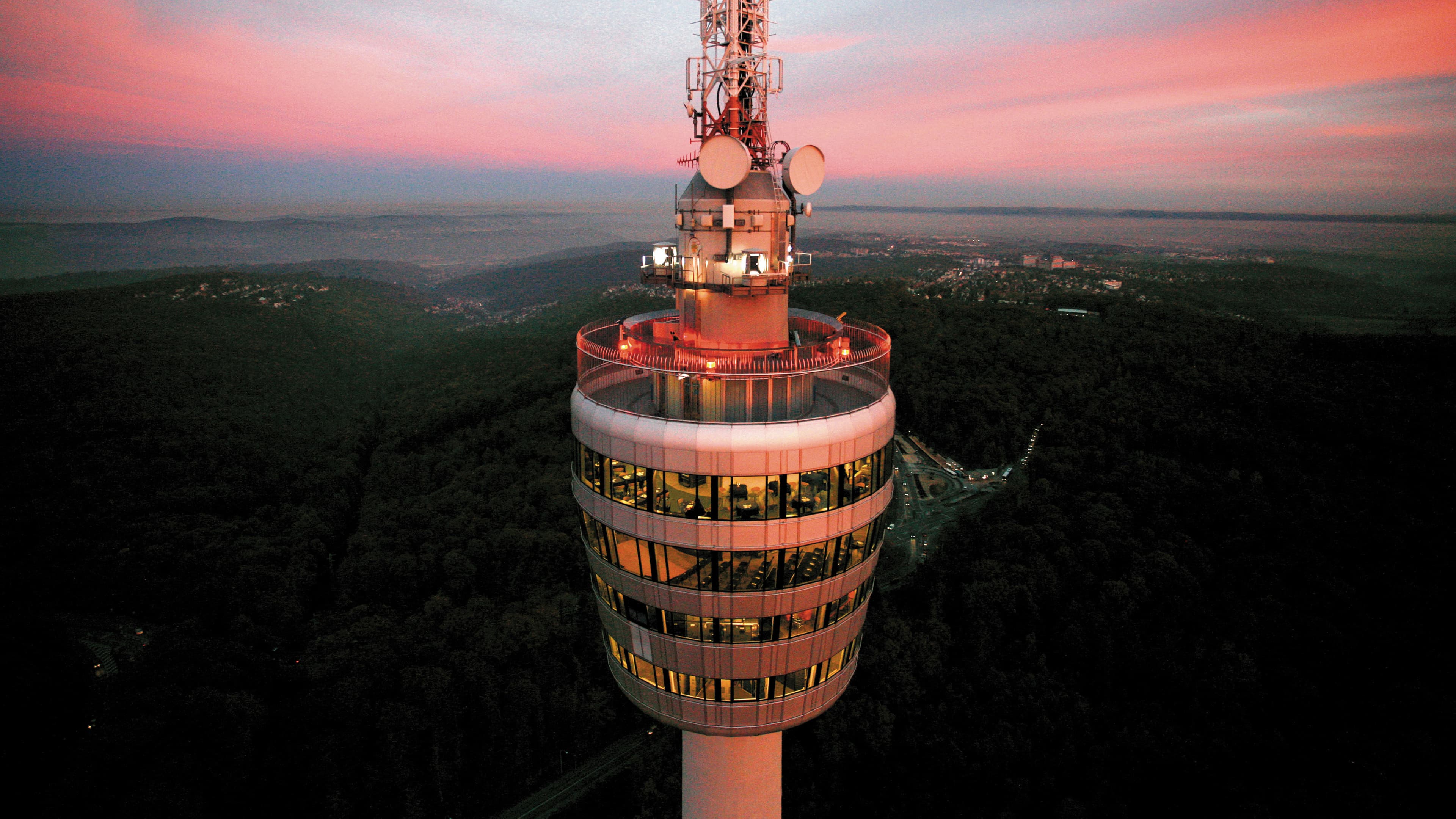 Der Fernsehturm Stuttgart bei Sonnenuntergang mit beleuchteter Aussichtsplattform und umliegendem Wald.
