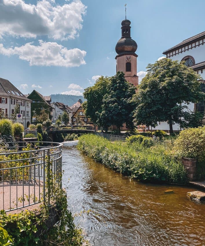 Auf dem Bild fließt ein Fluß durch eine malerische Stadt mit einem Zwiebelformkirchturm und Fachwerkhäsuern.