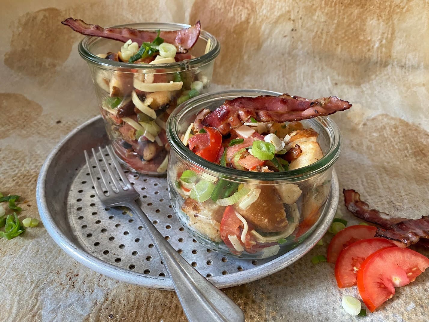 Auf einem Blechteller stehen zwei Gläser mit besonderen Salatkreationen mit Tomate, grünem Salat, Laugenbrötchen und Speck als Garnitur.