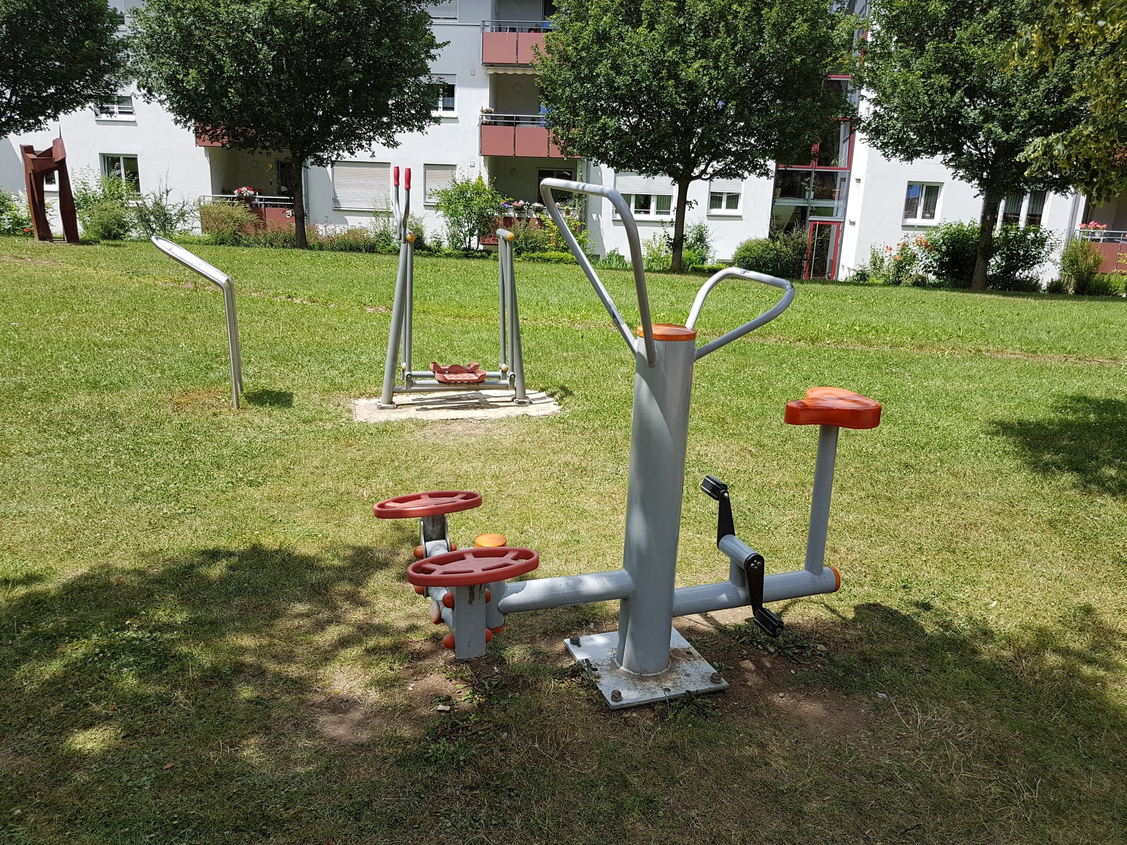 Zwei Outdoor-Trainingsgeräte stehen auf einer ebenen Fläche im Stadtgarten Hechingen. Sie sind aus Metall und Kunststoff gefertigt und fest im Boden verankert. Rundherum befinden sich Grünflächen, Bäume und Sitzbänke.